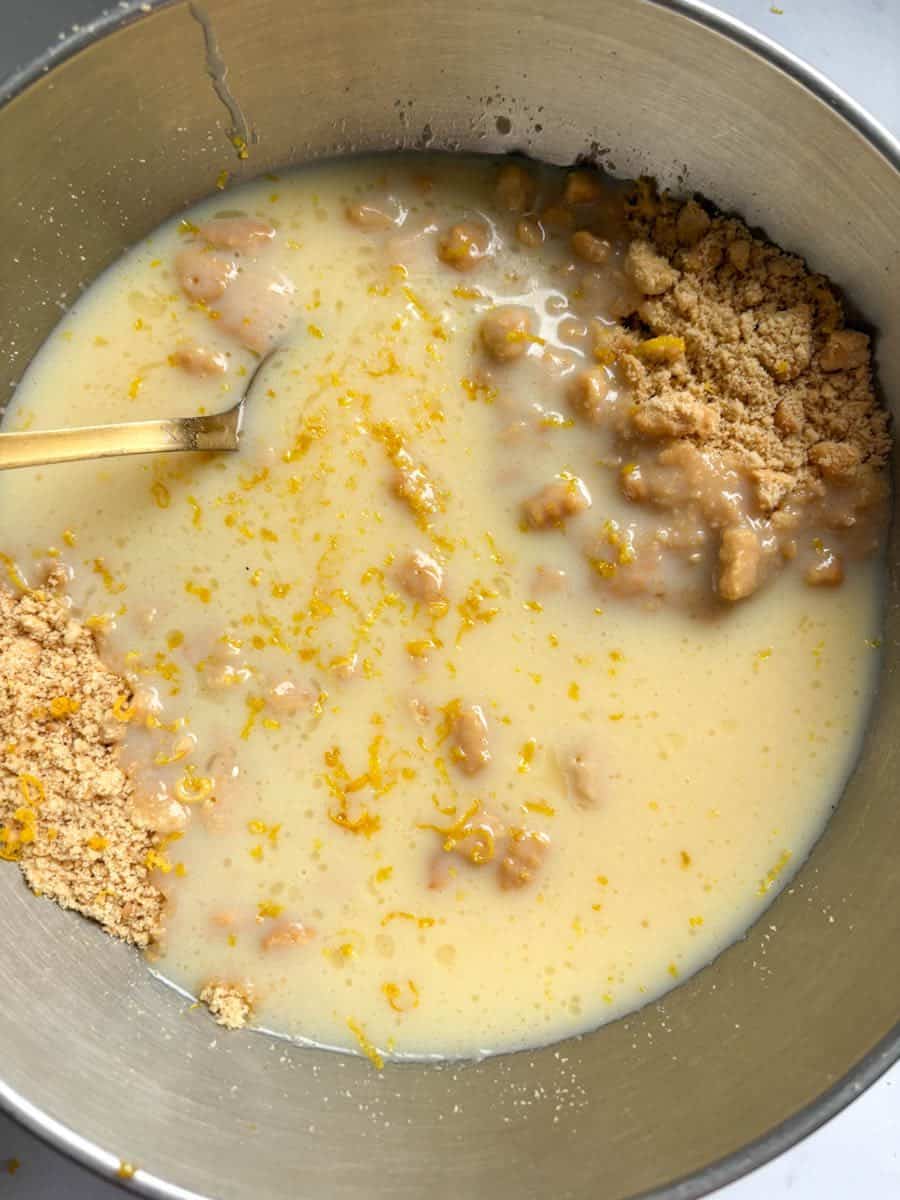 A silver bowl of condensed milk, crushed biscuits and lemon zest with a gold spoon.