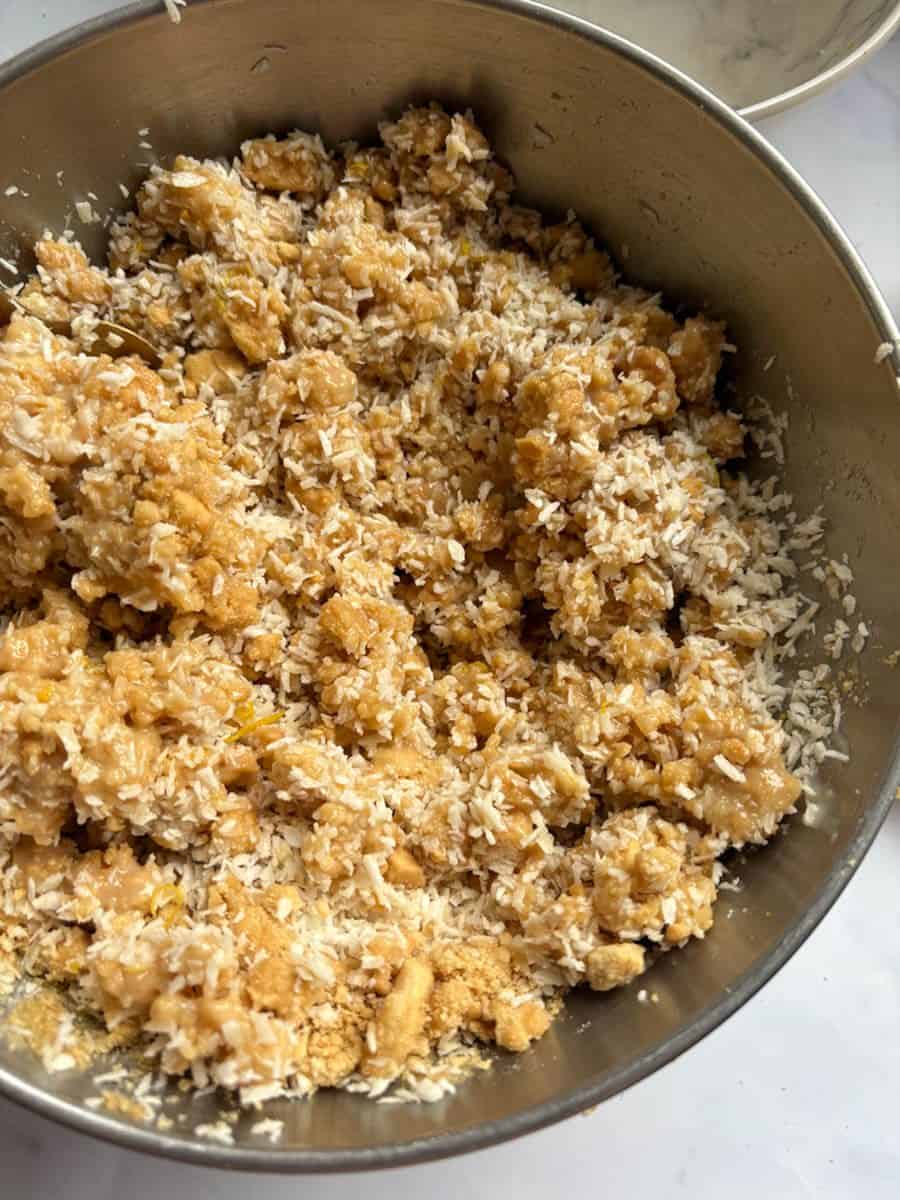 A silver bowl of crushed biscuits, melted butter, condensed milk, coconut and grated lemon zest.
