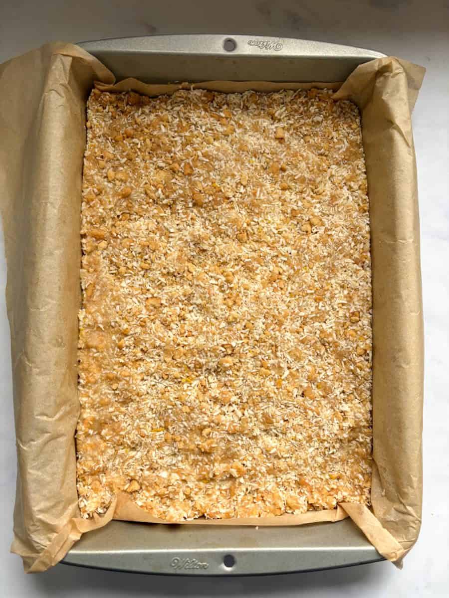 A lemon and coconut traybake with crushed biscuits in a lined baking tin.