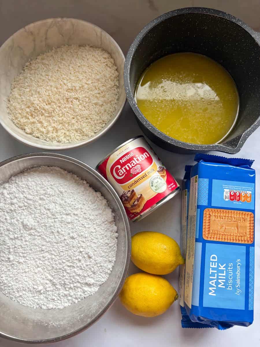Ingredients for making lemon fudge bars - a pan of melted butter, a packet of malted milk biscuits, two lemons, a tin of condensed milk, and bowls of icing sugar and desiccated coconut.