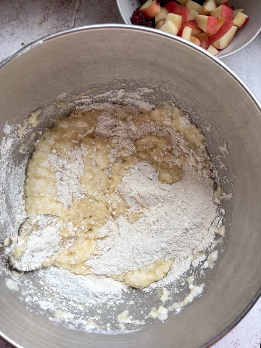 Flour being folded into a silver bowl of cake batter, with a bowl of diced peaches and blackberries in the background.