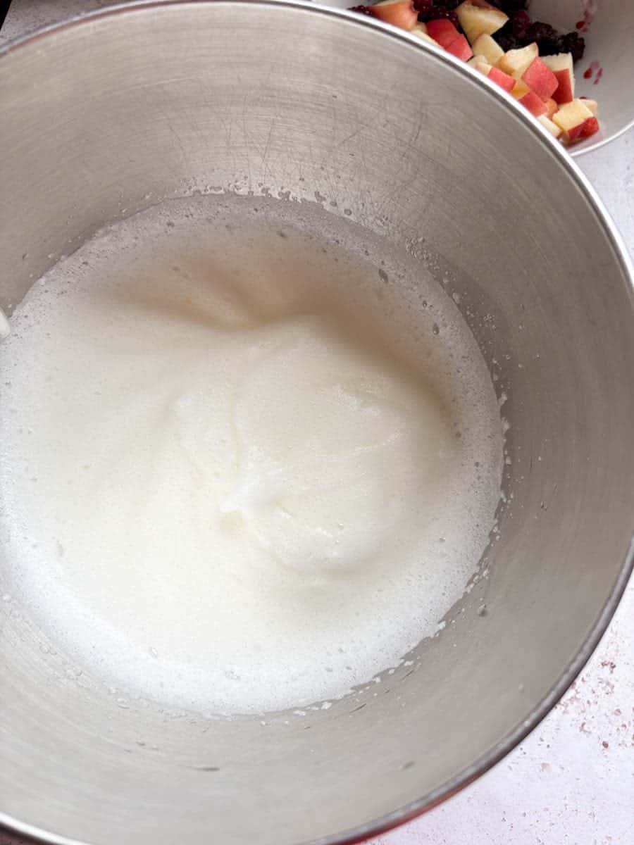 A large silver bowl of whisked egg whites. A white bowl of chopped peaches and blackberries can be partially seen.