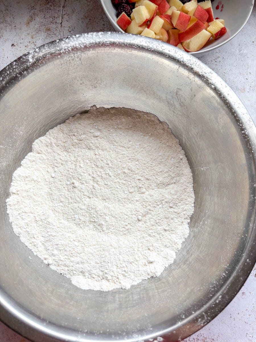 A large silver mixing bowl of icing sugar and a small bowl of chopped peaches and blackberries (partially seen).