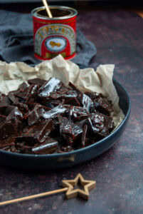 A black bowl of treacle toffee shards. The bowl is lined with a piece of brown baking parchment. A gold star decoration sits beside the bowl, and a red tin of Lyle's black treacle with a gold spoon.