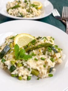 Two white bowls of Spring vegetable risotto with asparagus, diced courgettes, fresh peas, chives, a lemon slice and a sprig of fresh mint.