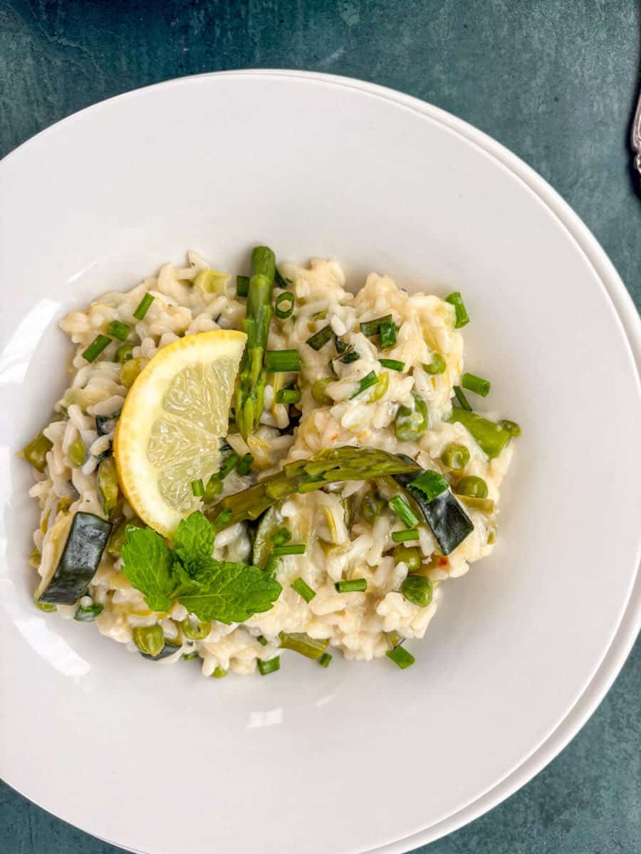 A white bowl of risotto with asparagus spears, diced courgettes, peas, chopped chives, a lemon slice and a sprig of fresh mint.